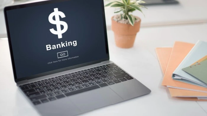 Open laptop displaying a digital banking interface with a dollar sign and 'Banking' text, placed on a desk with notebooks and a potted plant in the background, representing seamless money transfer services on the Mini Banking page of Ashoka Computer.